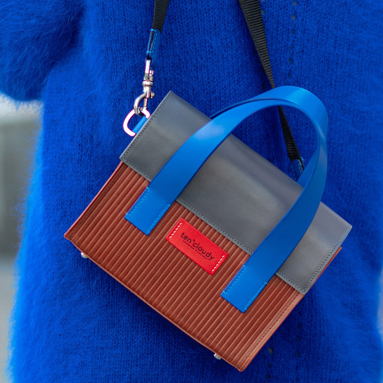 Small brown and gray handbag with a blue strap worn by a person in a blue coat.