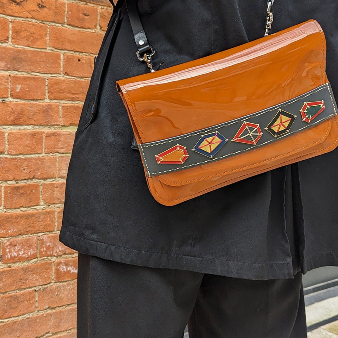 Person wearing a black coat with an orange handbag featuring geometric patterns against a brick wall.